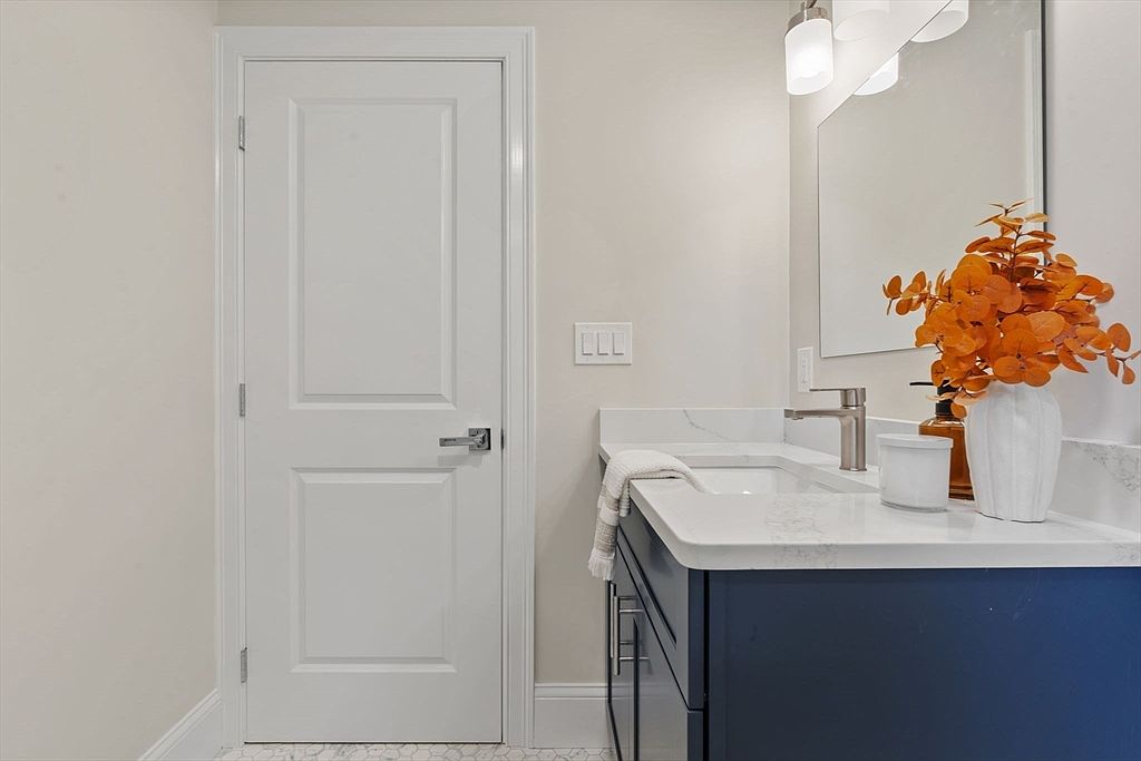 This is a well-lit bathroom featuring a navy blue vanity with a white countertop and a modern sink. A white door is visible to the left, and a decorative orange floral arrangement adds a pop of color. The overall impression is clean and stylish, suitable for a guest bathroom.