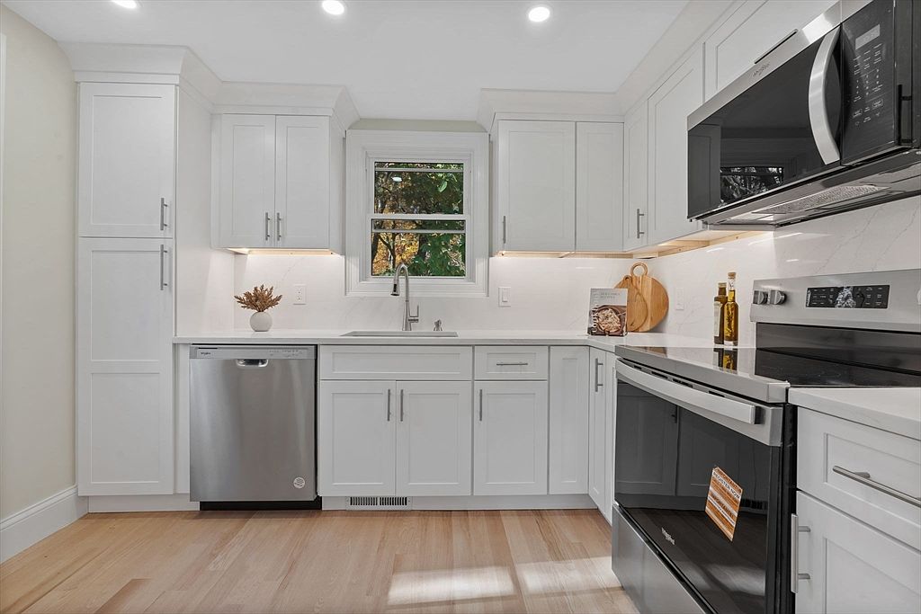 This is a well-lit kitchen featuring white cabinets, stainless steel appliances, and light hardwood floors. The kitchen includes a dishwasher, oven, microwave, and a sink under a window that provides natural light. The overall impression is clean, modern, and functional, making it an appealing space for cooking and meal preparation.