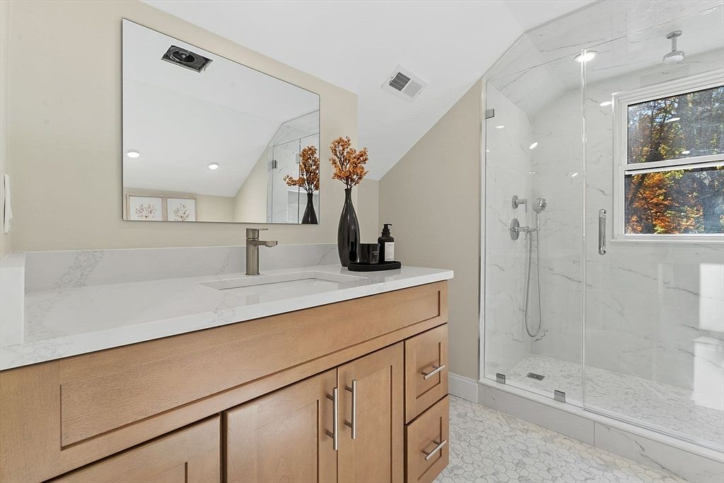 This is a bright and modern primary bathroom featuring a light wood vanity with a white countertop and a rectangular sink. A large mirror hangs above the vanity, reflecting the space and light. A glass-enclosed shower with marble-like tiling is visible to the right, adding a touch of luxury to the room.