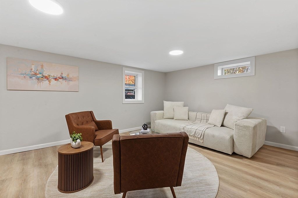 This is an interior shot of a living room featuring two brown armchairs, a light-colored sofa with throw pillows and a blanket, and a round side table with a plant. The room has light gray walls, wood-look flooring, and a round area rug, creating a cozy and inviting atmosphere. A piece of artwork hangs on the wall, and natural light enters through two small windows.