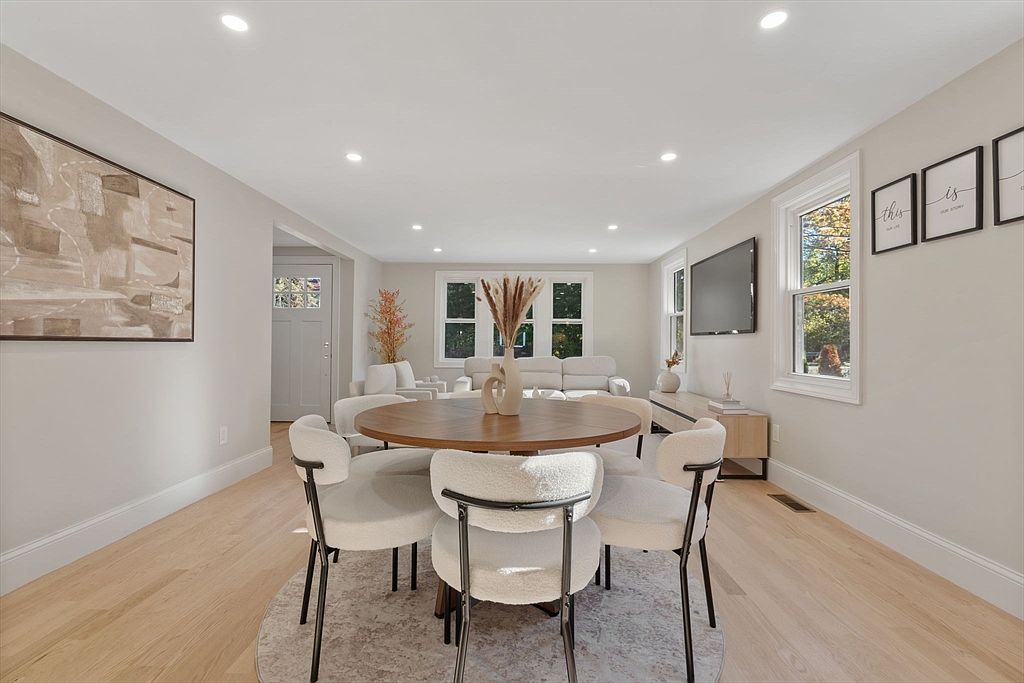 This interior shot showcases a dining area with a round wooden table surrounded by six modern chairs with white, textured upholstery and black metal legs. The room is brightly lit with recessed lighting and natural light from a nearby window, complemented by light hardwood floors and neutral wall colors. An abstract painting hangs on the wall, adding a touch of artistic flair to the space.