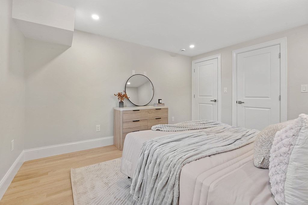 This is a well-lit bedroom featuring a light wood floor, a dresser with a round mirror above it, and a bed with a light-colored comforter and pillows. Two white doors are visible on the right side of the room. The room has a clean and modern aesthetic, creating a comfortable and inviting space.