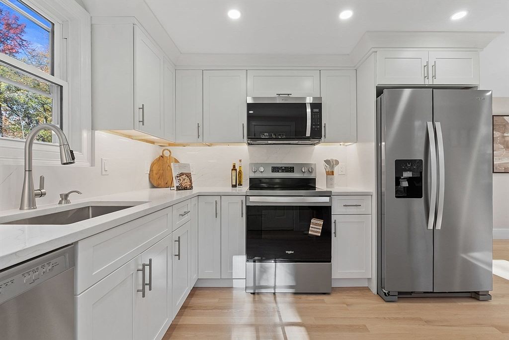 This is a well-lit kitchen featuring white cabinets with brushed metal hardware, stainless steel appliances, and light-colored countertops. The kitchen includes a stainless steel refrigerator, microwave, and oven. The flooring is light-colored wood, and a window provides natural light, creating a bright and inviting space.