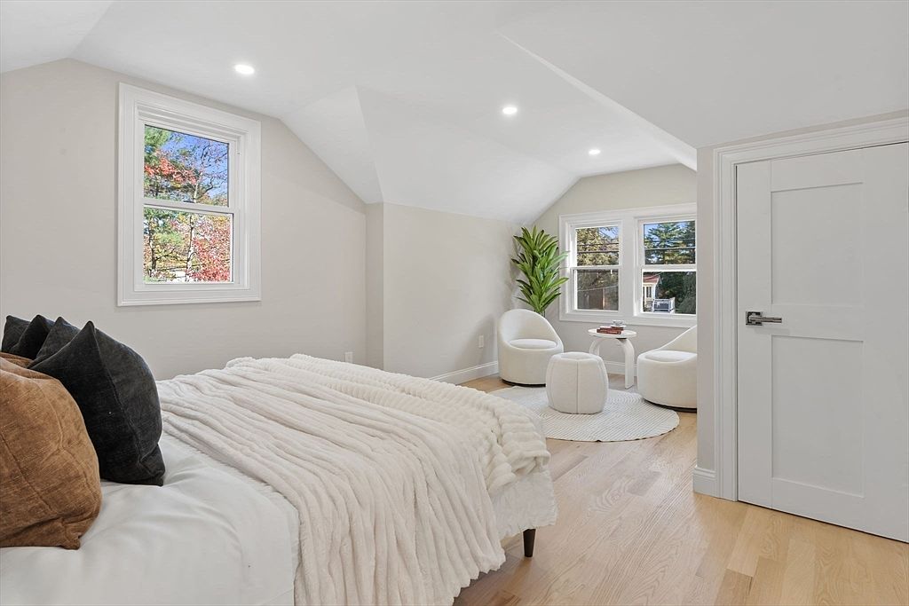 This is a bright and airy bedroom featuring a bed with a plush white blanket and decorative pillows. The room has two windows that let in natural light, illuminating the light hardwood floors and white walls. A small seating area with two chairs and a round rug adds a cozy touch to the space.