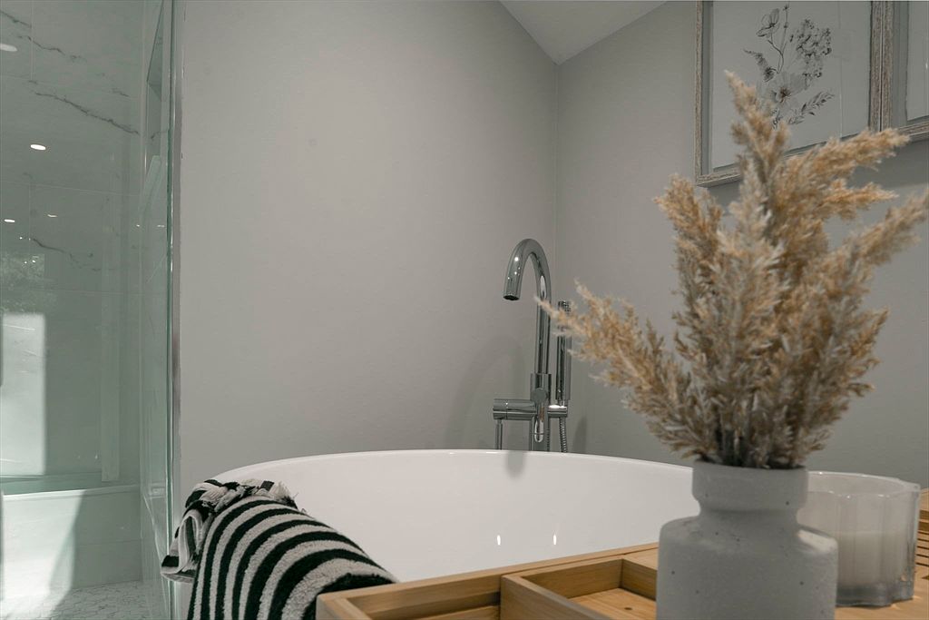 This is a close-up shot of a primary bathroom featuring a freestanding white bathtub with a modern chrome faucet. A wooden tray sits across the tub, holding a vase with dried floral arrangements and a candle. A striped towel is draped over the edge of the tub, adding a touch of texture and pattern to the serene and spa-like atmosphere.