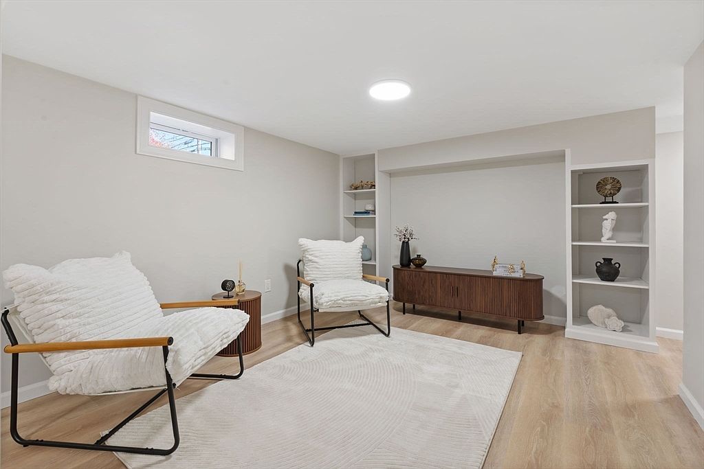 This is an interior shot of a living room featuring two modern chairs with white cushions, a light-colored rug, and a wooden cabinet against the wall. Built-in shelves flank the cabinet, and a small window provides natural light. The room has a minimalist and contemporary style, creating a cozy and inviting atmosphere.