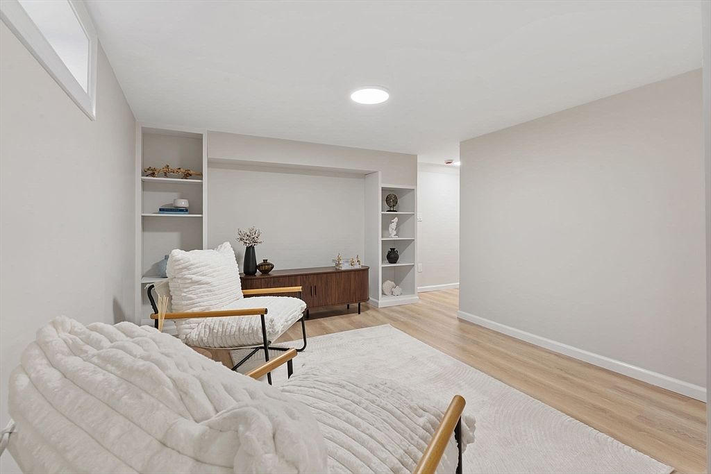 This is an interior shot of a living room featuring two armchairs with white cushions and wooden frames, placed on a light-colored rug. A mid-century modern style cabinet sits against the wall, flanked by built-in shelving units. The room is painted in neutral tones, creating a bright and airy atmosphere.