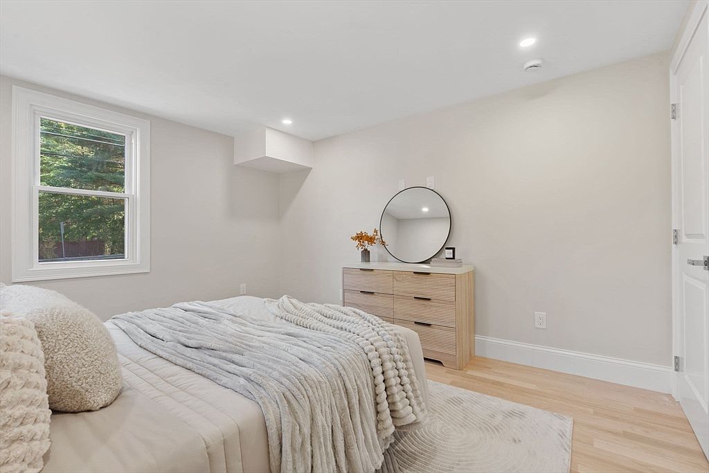 This is a bright and airy bedroom featuring a bed with a light-colored duvet and decorative pillows. A wooden dresser with a round mirror sits against the wall, complemented by light hardwood floors and white trim. The room has a clean and minimalist aesthetic, creating a calming and inviting space.