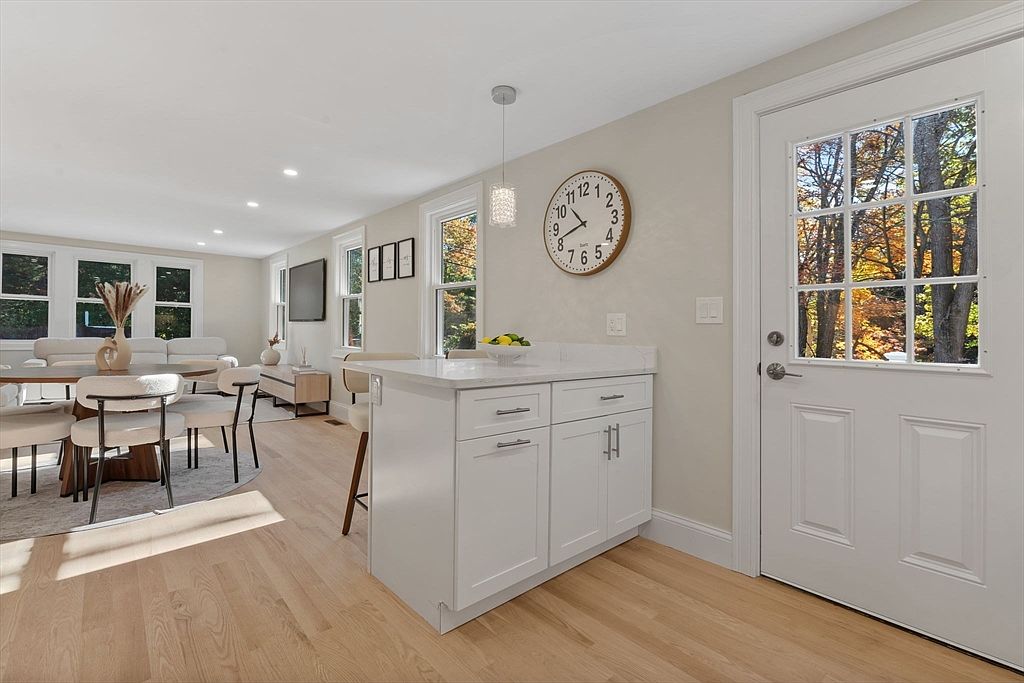 This interior shot showcases a bright and airy living space that seamlessly blends into a dining area and kitchen. The room features light hardwood floors, a modern dining set with white chairs, and a kitchen island with white cabinetry. A door with glass panes offers a view of the outdoors, enhancing the natural light in the space.