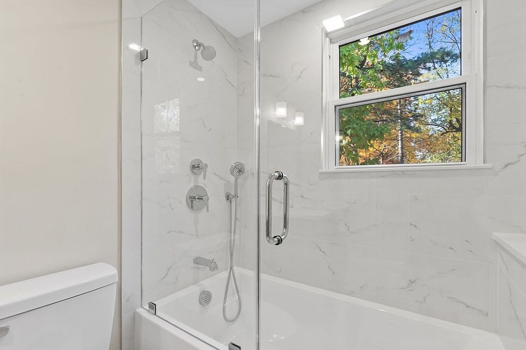 This is a bright and modern bathroom featuring white marble-style tiling throughout the shower and walls. A glass shower door encloses the tub/shower combination, which includes a rainfall showerhead and handheld sprayer. A window provides natural light and a view of the trees outside, enhancing the spa-like atmosphere.