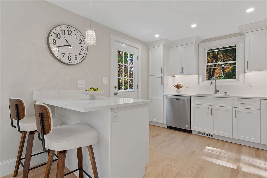 This is a bright and modern kitchen featuring white cabinetry, stainless steel appliances, and light hardwood floors. A breakfast bar with stylish stools provides seating, while a large clock adds a decorative touch. Natural light streams in through a window above the sink, creating a welcoming atmosphere.