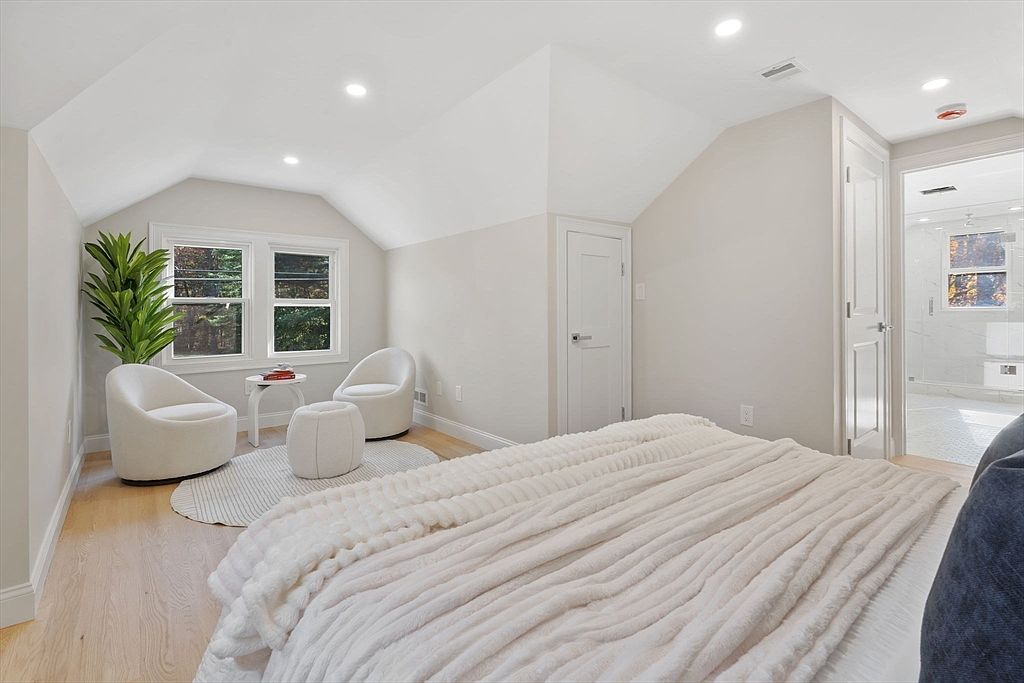 This is a bright and airy primary bedroom featuring a neutral color palette. The room includes a seating area with two white chairs and a small table near a window, a large bed with a plush white blanket, and an ensuite bathroom visible through an open doorway. The hardwood floors and recessed lighting add to the room's modern and inviting atmosphere.