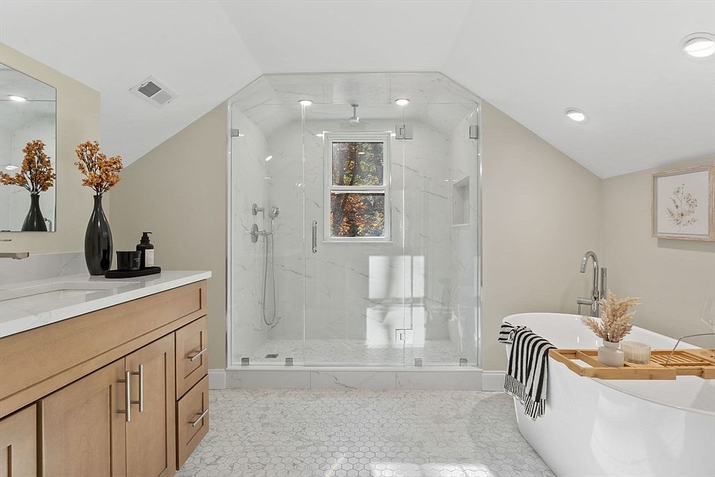This is a bright and airy primary bathroom featuring a glass-enclosed shower with marble-like tile, a freestanding bathtub with a bamboo tray, and a vanity with a light wood finish and white countertop. The floor is covered in a small hexagon tile pattern, and the room is well-lit with recessed lighting and natural light from a window in the shower. The overall impression is clean, modern, and luxurious.