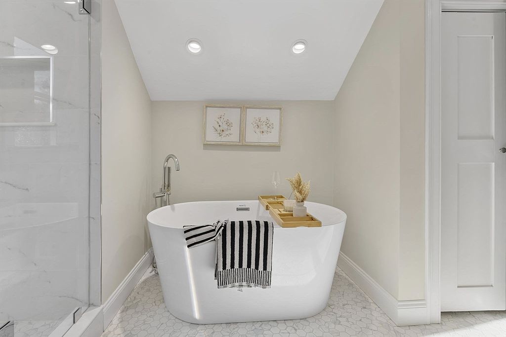 This is a bright and airy primary bathroom featuring a freestanding white bathtub as the focal point. The room is decorated with neutral tones, including light beige walls and white trim, complemented by two framed botanical prints above the tub. The bathroom also includes a glass-enclosed shower and a white door, creating a clean and relaxing atmosphere.
