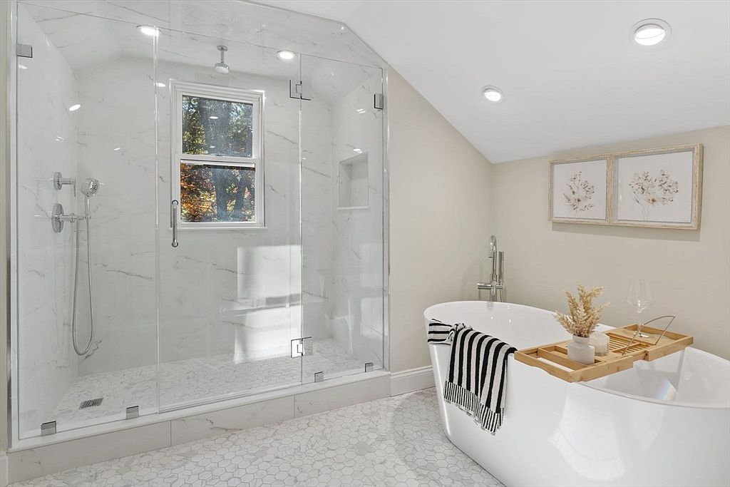 This is a bright and luxurious primary bathroom featuring a glass-enclosed shower with marble-like tile and a window. A freestanding white bathtub is accessorized with a bamboo tray, decorative items, and a striped towel. The floor is tiled with a small hexagonal pattern, and the walls are painted in a neutral tone, creating a serene and spa-like atmosphere.