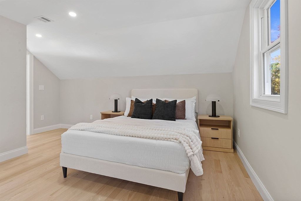 This is a bright and airy bedroom featuring a bed with a white frame and bedding, complemented by dark pillows and a textured throw. Two matching nightstands with lamps flank the bed, and a window provides natural light, highlighting the light wood flooring. The room has a clean and minimalist aesthetic, perfect for a guest room.