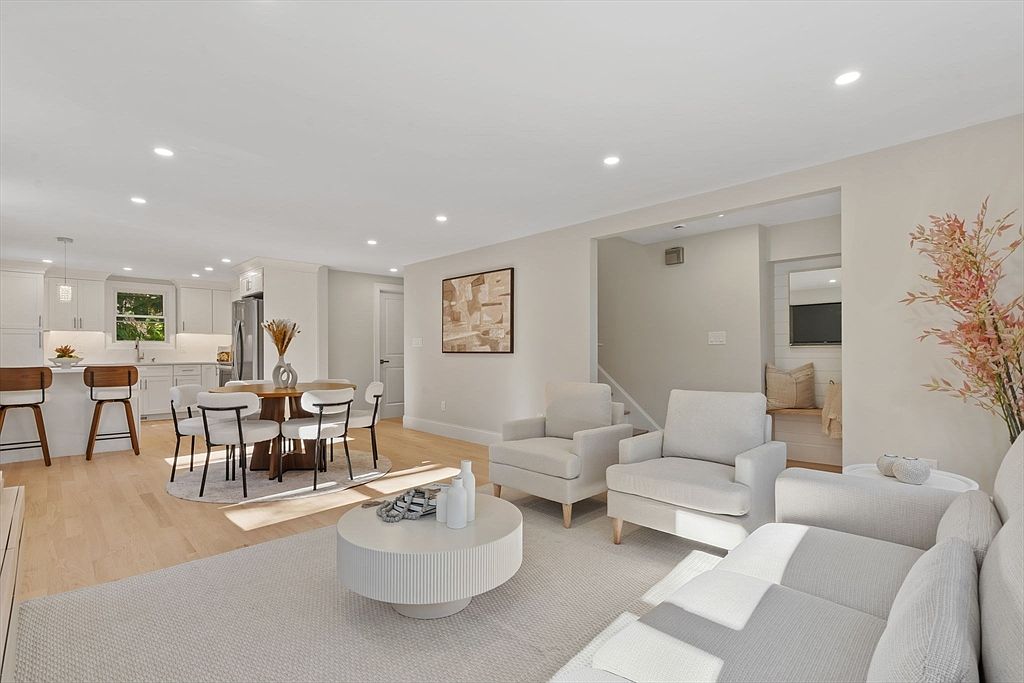 This is an interior shot of a combined living room, dining area, and kitchen. The living room features a neutral color palette with white sofas, armchairs, and a round coffee table, all set on a light-colored rug. The dining area includes a round table with modern chairs, and the kitchen has white cabinets and stainless steel appliances, creating a cohesive and bright living space.
