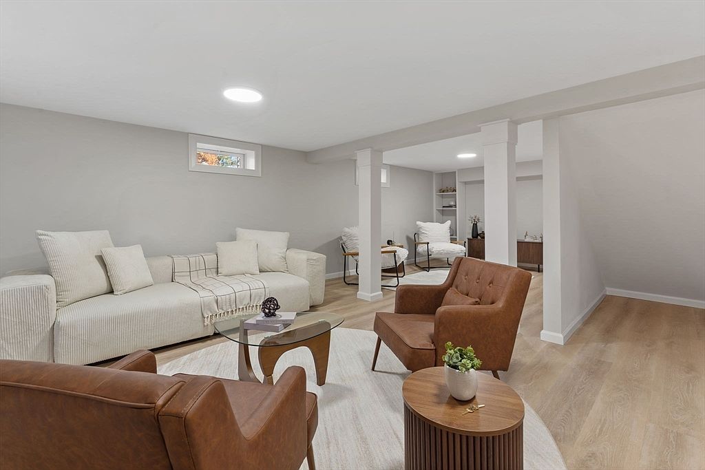 This is a well-staged living room featuring a neutral color palette with a large, comfortable sofa adorned with throw pillows and a blanket. Two brown leather armchairs face the sofa, creating a cozy conversation area. A round area rug anchors the space, and a glass coffee table and wooden side table add functional elegance, making the room feel inviting and modern.