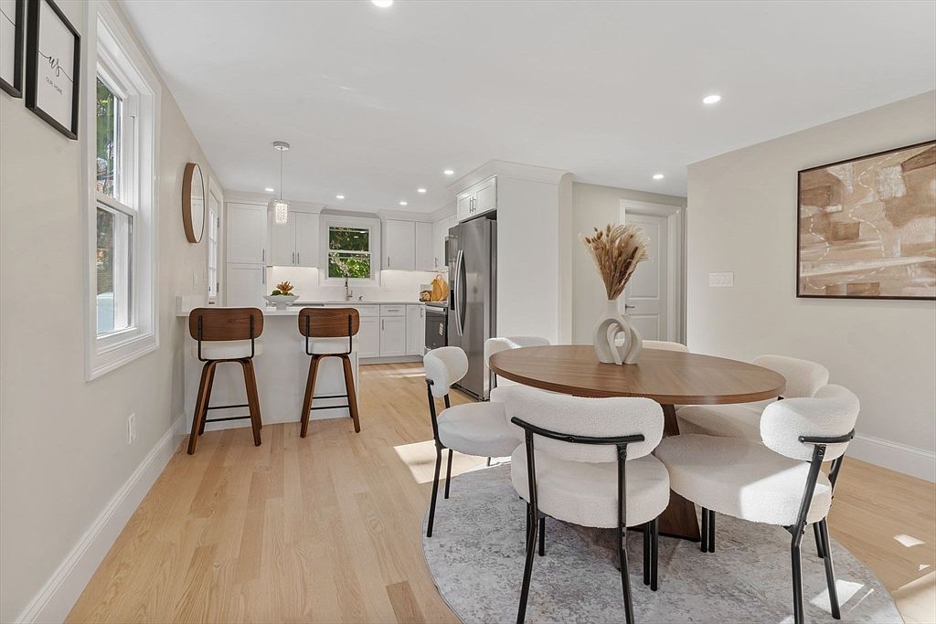 This interior shot showcases a dining area adjacent to a modern kitchen. The dining space features a round wooden table surrounded by six stylish chairs with white, textured upholstery and black frames, set upon a patterned rug. The kitchen is visible in the background, featuring white cabinets and stainless steel appliances, creating a cohesive and inviting living space.