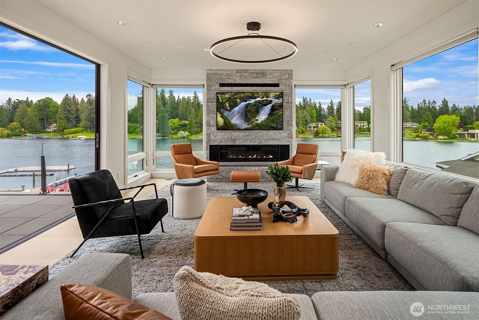 This sophisticated, modern living room features an expansive open-concept design with floor-to-ceiling windows that offer panoramic views of a serene lake and lush greenery. The space is anchored by a striking stone fireplace with a built-in television, complemented by a large, light-wood coffee table and a plush grey sectional sofa. The room exudes a luxurious, tranquil atmosphere, enhanced by a contemporary circular chandelier and warm, inviting leather accent chairs.