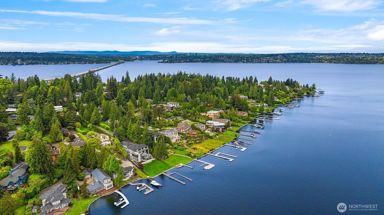 This high-angle aerial view captures a luxurious waterfront neighborhood nestled along a lush, tree-lined shoreline. The scene features several upscale homes with private docks extending into the expansive, calm blue lake, with a distant bridge spanning the horizon under a bright, cloudy sky. The perspective emphasizes the serene, exclusive nature of the property and its prime location on the water.