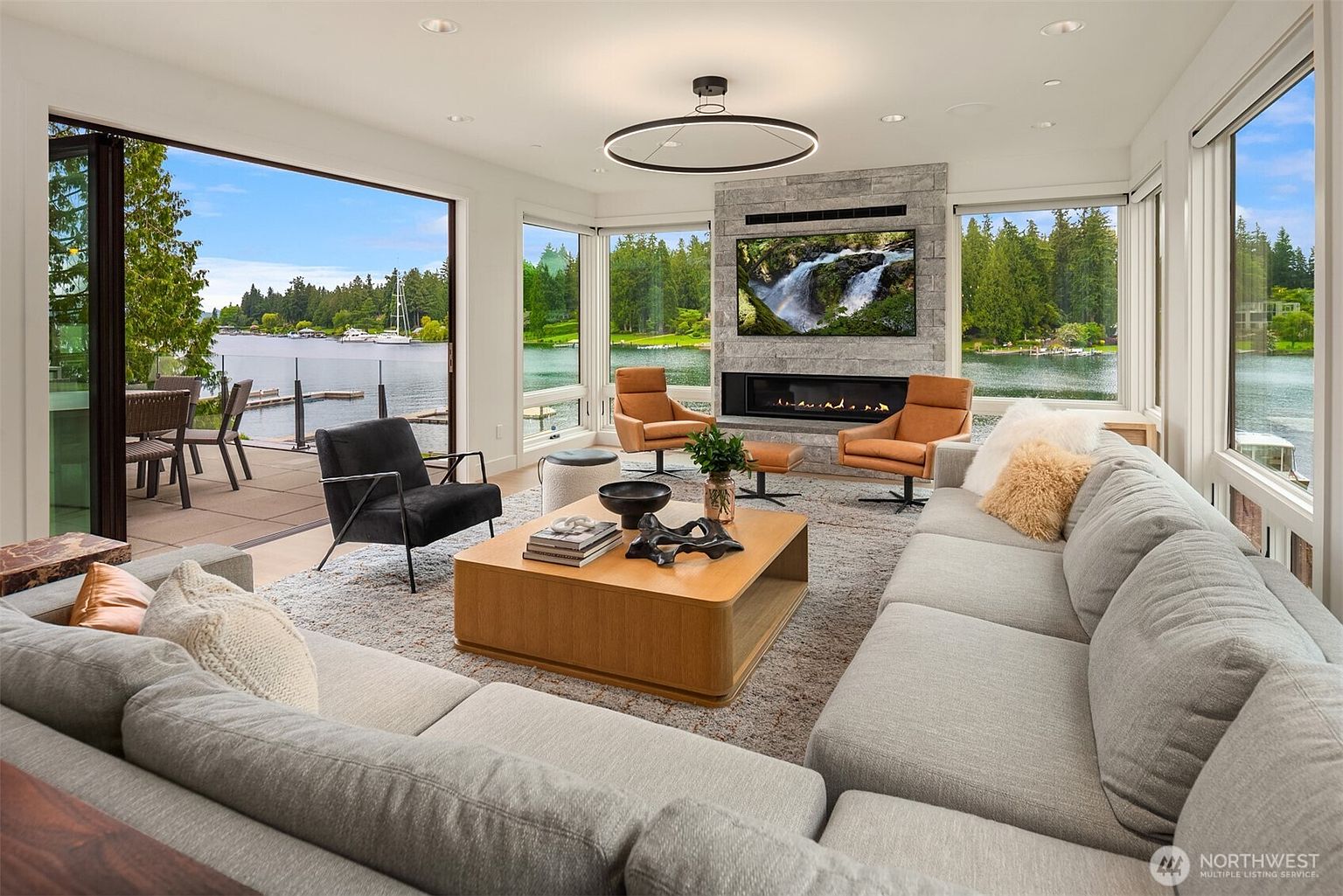 This sophisticated, open-concept living room features a large, plush grey sectional sofa and a central wooden coffee table, all centered around a striking stone fireplace with an integrated television. Floor-to-ceiling glass walls provide expansive, panoramic views of a serene lake and lush greenery, seamlessly blending indoor comfort with the natural landscape. The space is accented by modern leather armchairs and a minimalist circular chandelier, creating an atmosphere of refined, contemporary luxury.