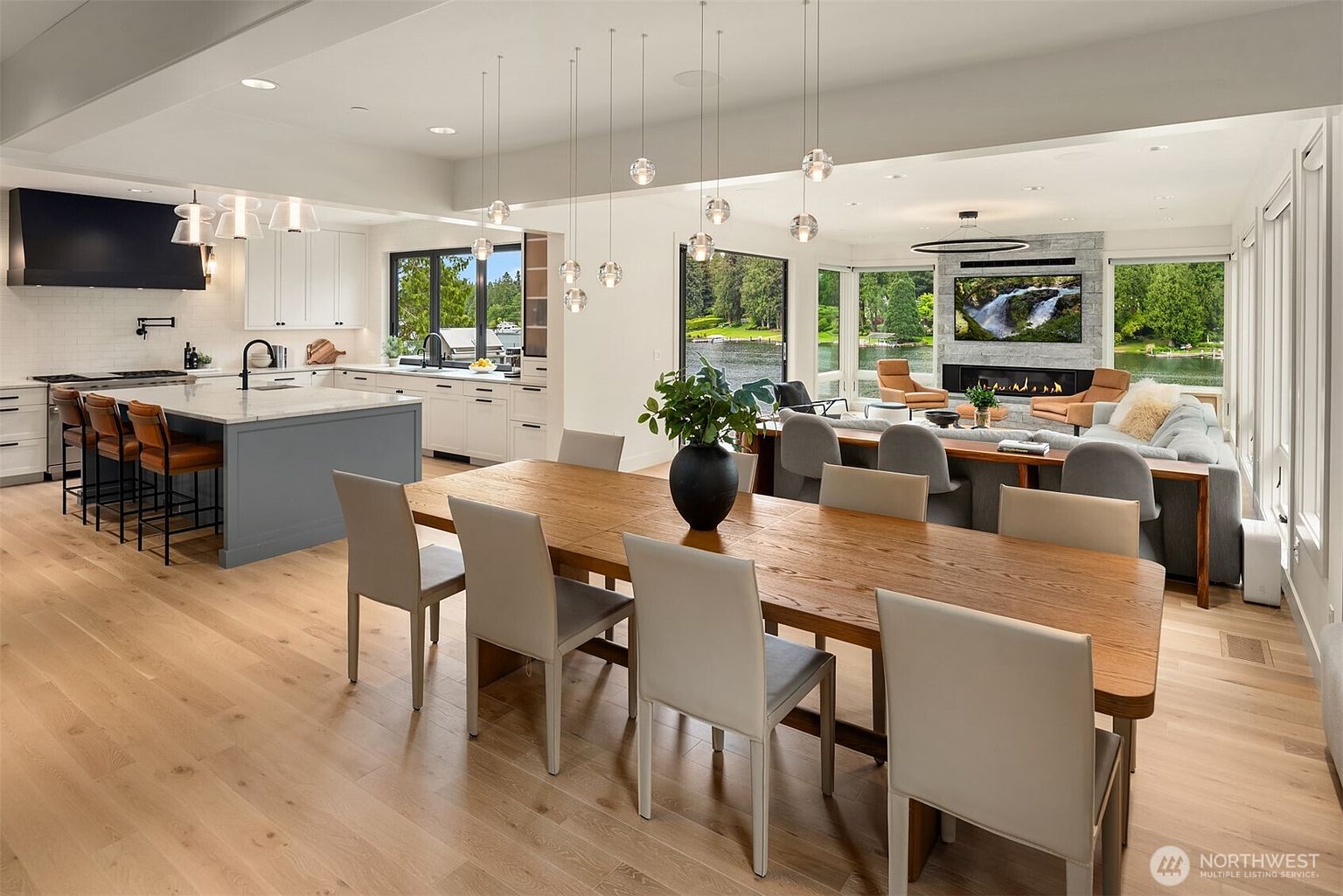 This expansive, open-concept living space seamlessly blends a modern kitchen, dining area, and living room, all bathed in natural light from floor-to-ceiling windows overlooking a serene lake. The kitchen features a large gray island with bar seating, white cabinetry, and high-end finishes, while the dining area is anchored by a substantial wooden table. The overall aesthetic is sophisticated and airy, emphasizing a luxurious, indoor-outdoor lifestyle with a contemporary fireplace serving as a focal point in the living area.