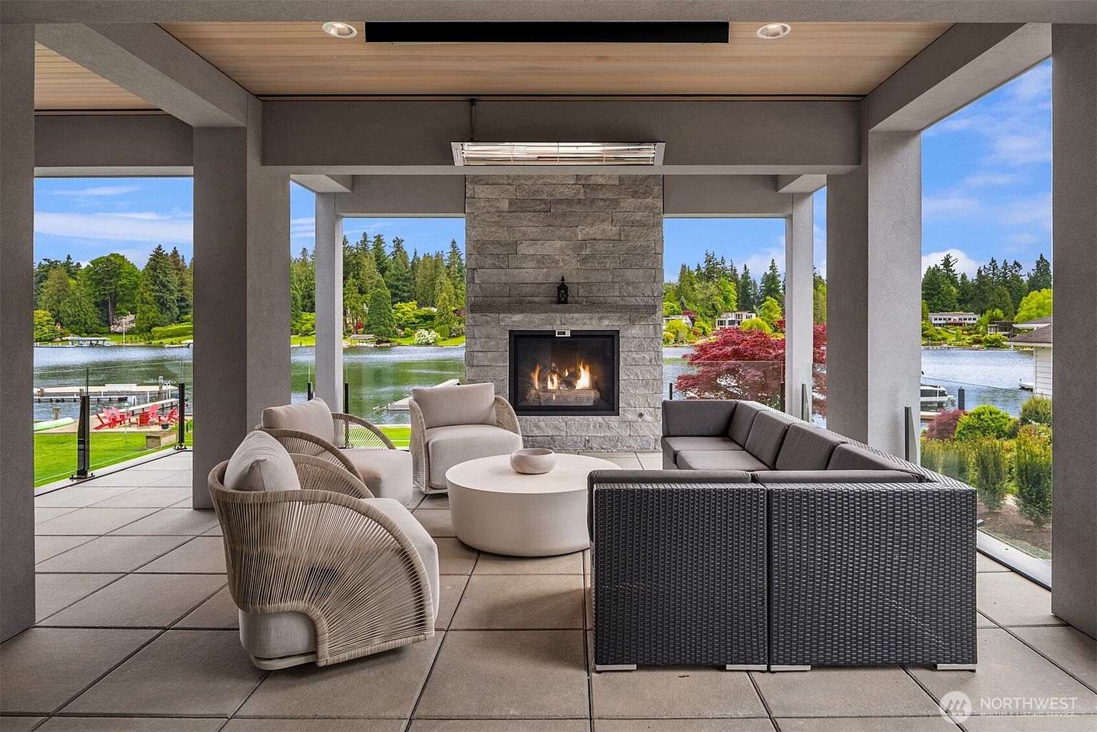This luxurious covered outdoor patio features a modern stone fireplace as its centerpiece, flanked by expansive glass railings that offer panoramic views of a serene lake and lush greenery. The space is furnished with a sophisticated grey sectional sofa, two woven accent chairs, and a minimalist round coffee table, creating an inviting atmosphere for relaxation. The high-end design is complemented by a warm wood-paneled ceiling and recessed lighting, emphasizing a seamless blend of indoor comfort and outdoor beauty.