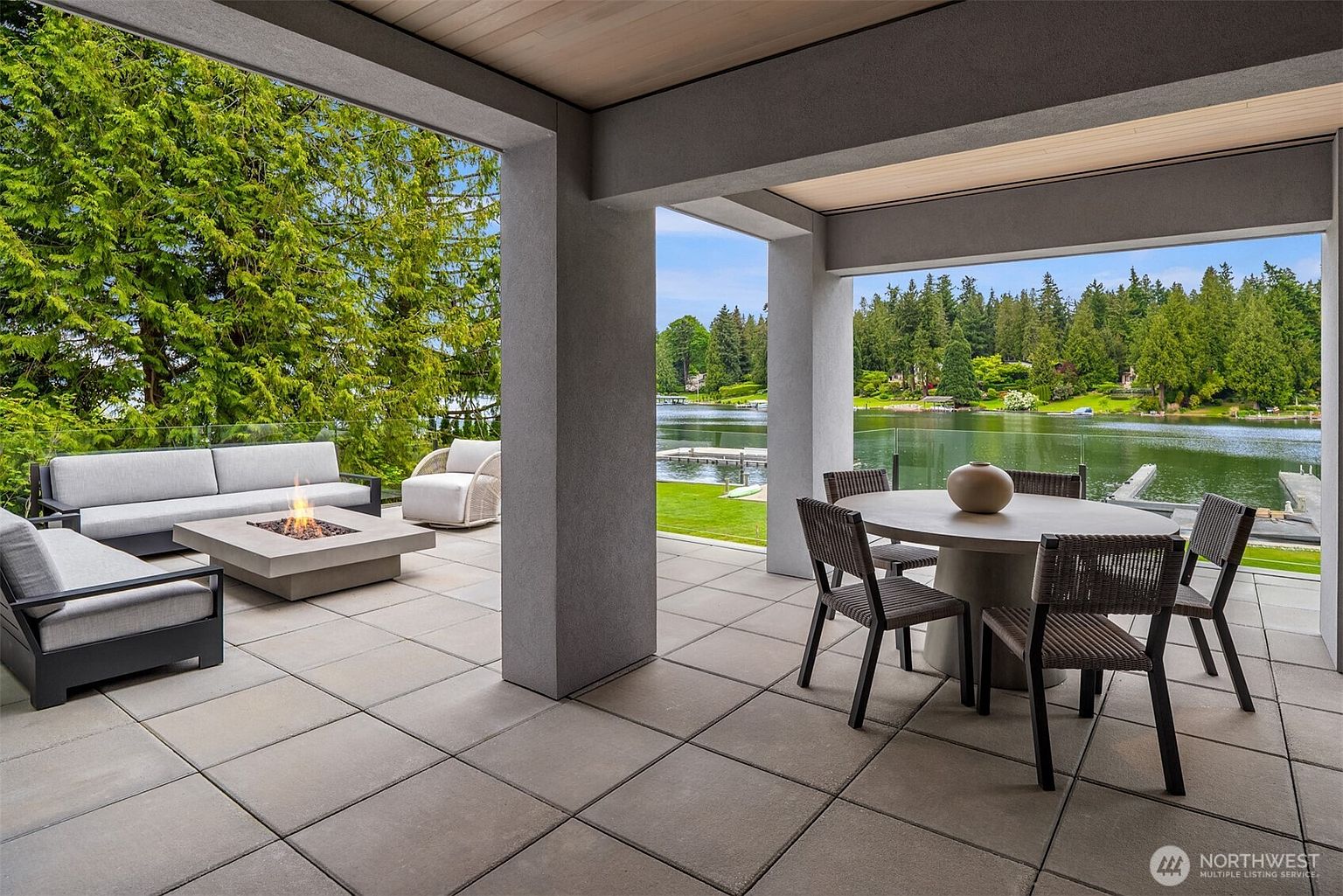 This luxurious covered patio offers a seamless transition between indoor comfort and outdoor living, featuring a modern concrete fire pit area and a sophisticated dining space. The space is framed by large structural pillars and overlooks a serene lake, with glass railings ensuring an unobstructed view of the lush, tree-lined waterfront. The neutral color palette and clean lines create a tranquil, high-end atmosphere perfect for relaxation or entertaining.