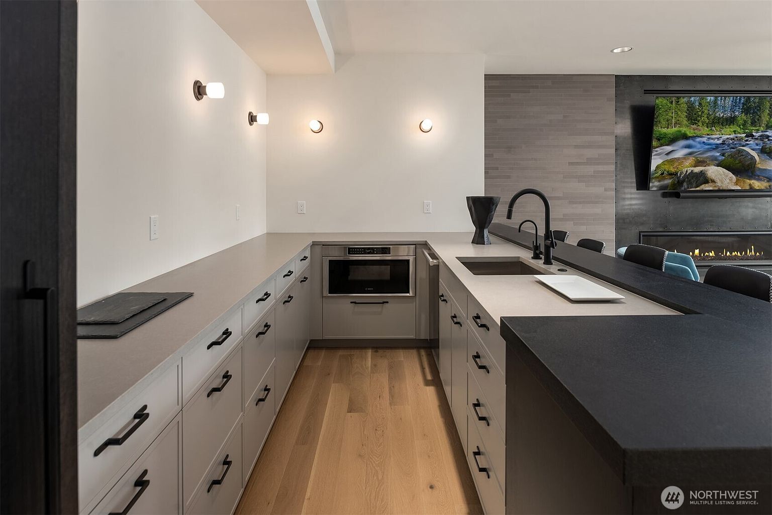 This modern, U-shaped kitchen features sleek light-gray cabinetry paired with contrasting dark countertops and warm hardwood flooring. The space is equipped with a built-in microwave, a minimalist faucet, and contemporary wall-mounted lighting, creating a clean and sophisticated aesthetic. The perspective looks directly into the kitchen area, highlighting the seamless transition between the workspace and the adjacent living area with a fireplace.