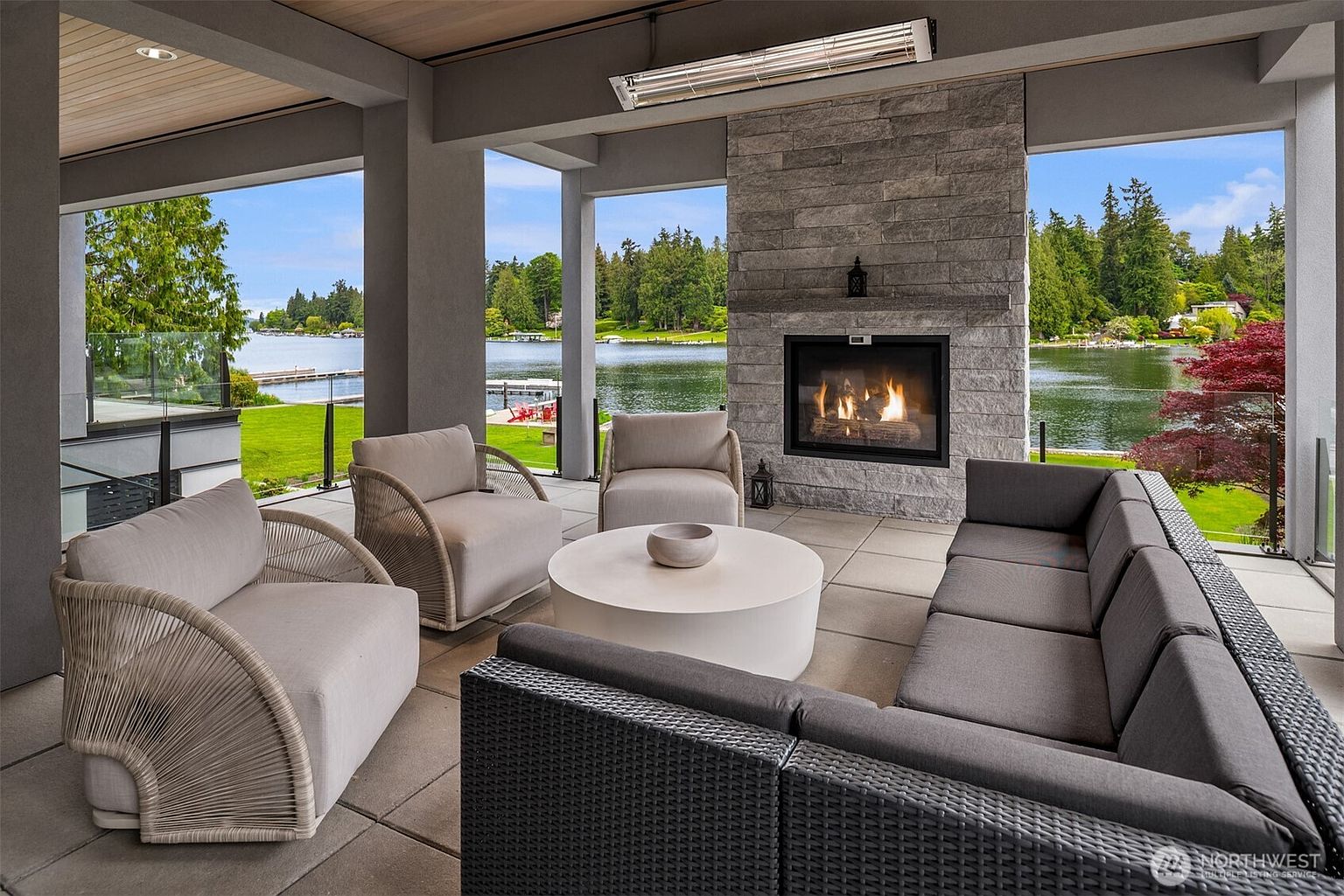 This luxurious covered patio offers a sophisticated outdoor living space, featuring a large stone fireplace as the focal point and comfortable, modern lounge seating. The open-air design seamlessly connects the interior comfort to the stunning waterfront views of the lake and surrounding greenery. The space is perfectly curated for relaxation and entertaining, blending high-end materials with a serene, natural backdrop.