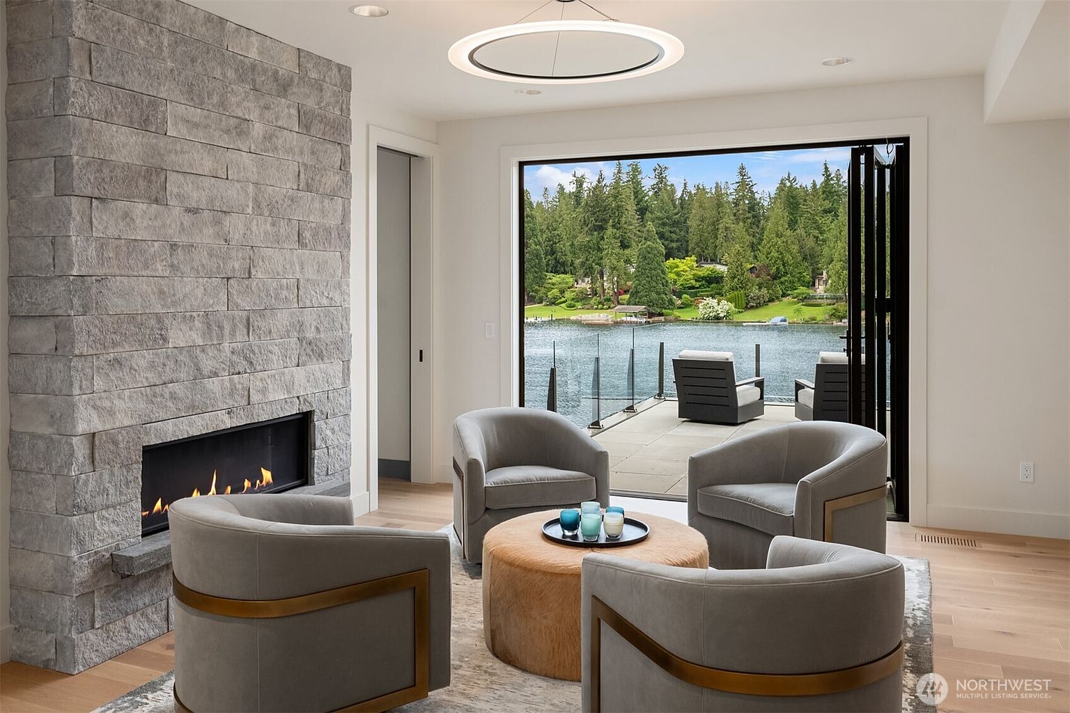 This sophisticated living room features a striking floor-to-ceiling stone fireplace and a circular modern chandelier, creating a luxurious and cozy atmosphere. The space is anchored by four plush, curved armchairs surrounding a textured ottoman, all set against a backdrop of expansive folding glass doors that seamlessly blend the interior with a serene lakefront patio. The overall aesthetic is contemporary and elegant, emphasizing a strong connection to the natural landscape outside.