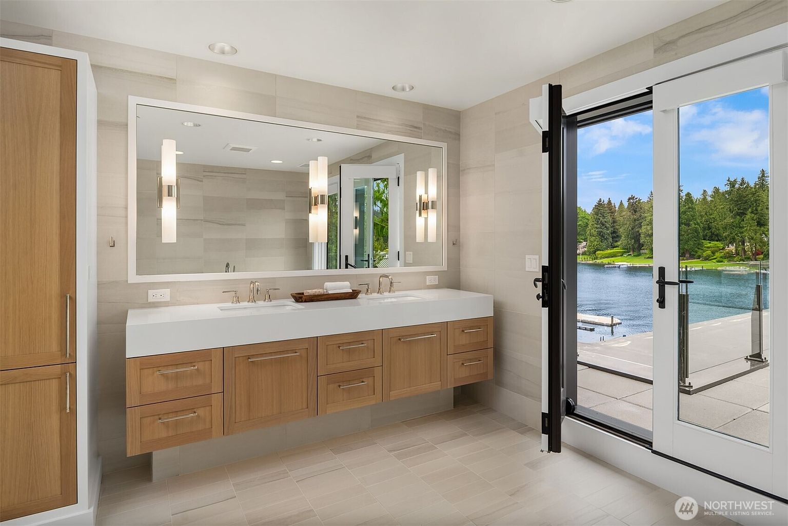 This luxurious primary bathroom features a sleek, floating double vanity with light wood cabinetry and a crisp white countertop. A large, expansive mirror reflects the room, while floor-to-ceiling glass doors open directly to a stunning waterfront view, blending indoor elegance with natural beauty. The neutral, large-format tile flooring and walls create a serene, spa-like atmosphere perfect for a high-end residence.