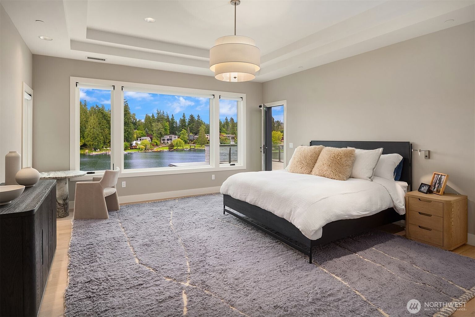 This spacious primary bedroom features a contemporary design with a large, plush area rug and a king-sized bed as the focal point. A wall of expansive windows provides a stunning, panoramic view of the lake and surrounding greenery, while a small marble-topped desk area offers a serene workspace. The room is bathed in natural light, creating an airy and luxurious atmosphere perfect for a waterfront retreat.