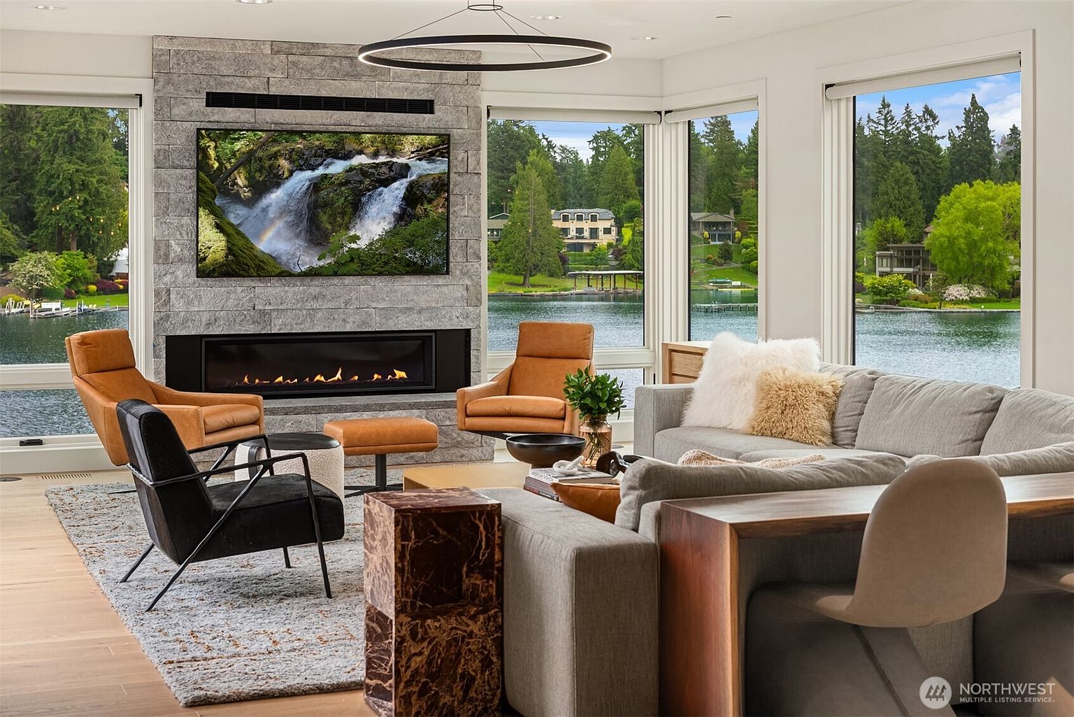 This sophisticated living room features a striking floor-to-ceiling stone fireplace with a built-in linear hearth and a large wall-mounted television. Expansive floor-to-ceiling windows frame a serene, panoramic view of a lake and lush forest, bathing the room in natural light. The space is furnished with a plush grey sectional, a modern black accent chair, and warm leather armchairs, creating a luxurious and inviting atmosphere.