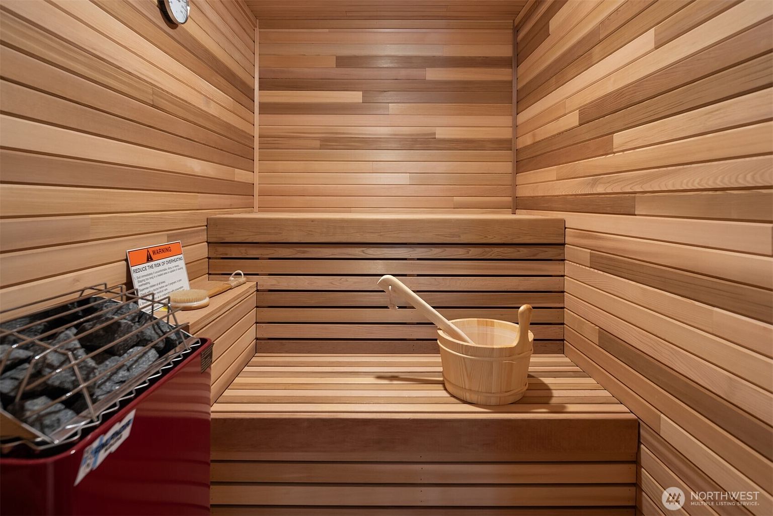 This image showcases a compact, wood-paneled sauna featuring horizontal cedar slats and a built-in bench. A red electric heater filled with dark stones sits in the foreground, accompanied by a wooden bucket and ladle, creating a relaxing and authentic spa-like atmosphere. The warm, natural wood tones and clean lines provide a sense of comfort and luxury, making it an appealing wellness feature for a home.