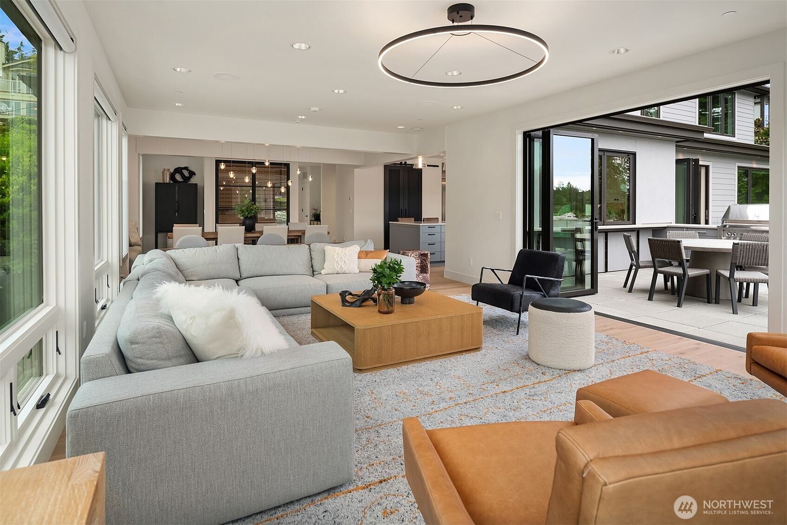 This spacious, open-concept living room features a large, light-gray sectional sofa, a central wooden coffee table, and two tan leather armchairs, creating a warm and inviting atmosphere. The room seamlessly transitions to an outdoor patio through expansive, black-framed folding glass doors, highlighting a modern indoor-outdoor lifestyle. A striking, large circular pendant light serves as a focal point above the seating area, while the neutral color palette and clean lines emphasize a contemporary, high-end aesthetic.