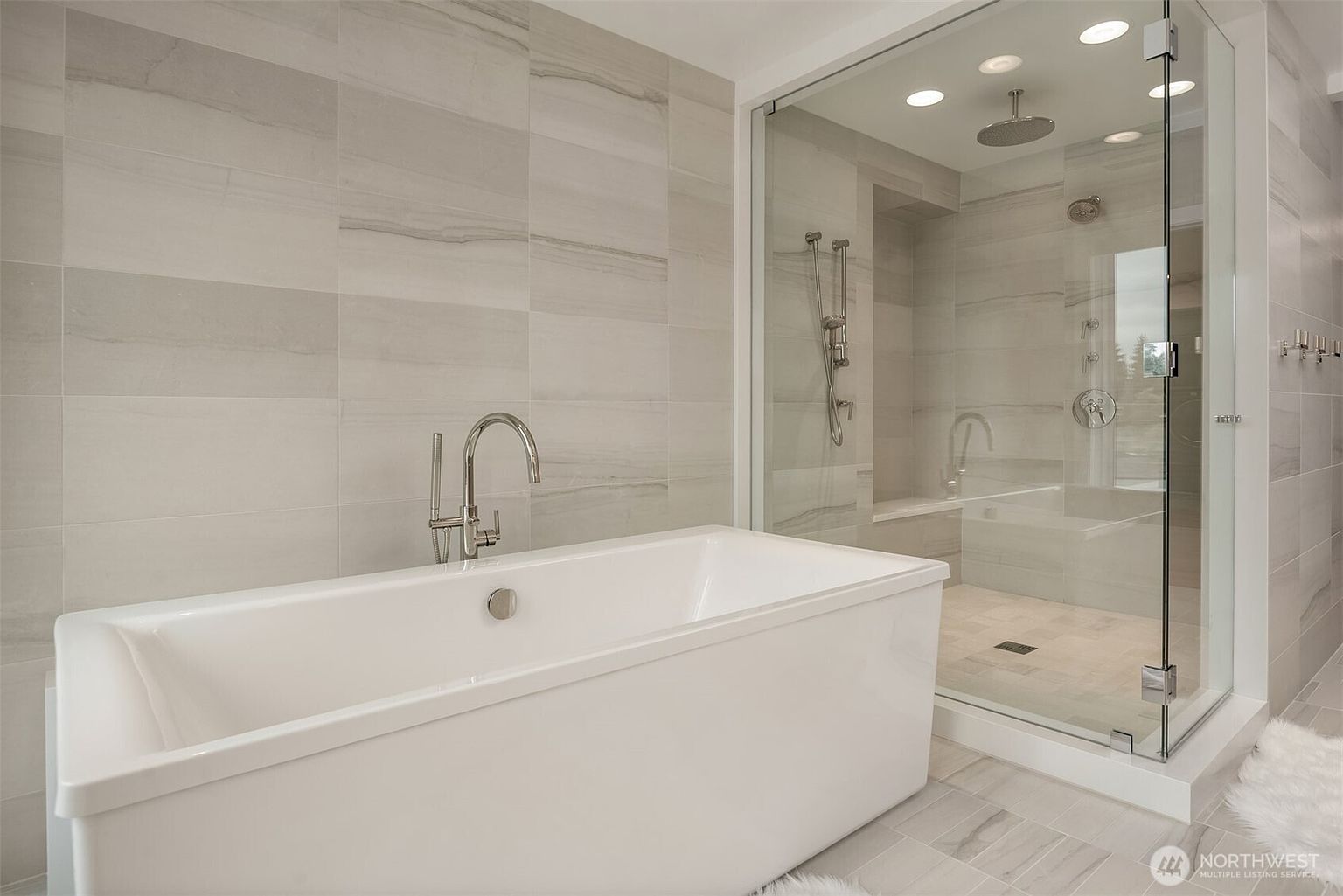 This modern primary bathroom features a sleek, white freestanding soaking tub paired with a contemporary chrome floor-mounted faucet. Adjacent to the tub is a spacious walk-in glass-enclosed shower with light-toned, large-format tile walls that create a bright and airy atmosphere. The minimalist design and neutral color palette offer a clean, spa-like aesthetic perfect for a luxury home.
