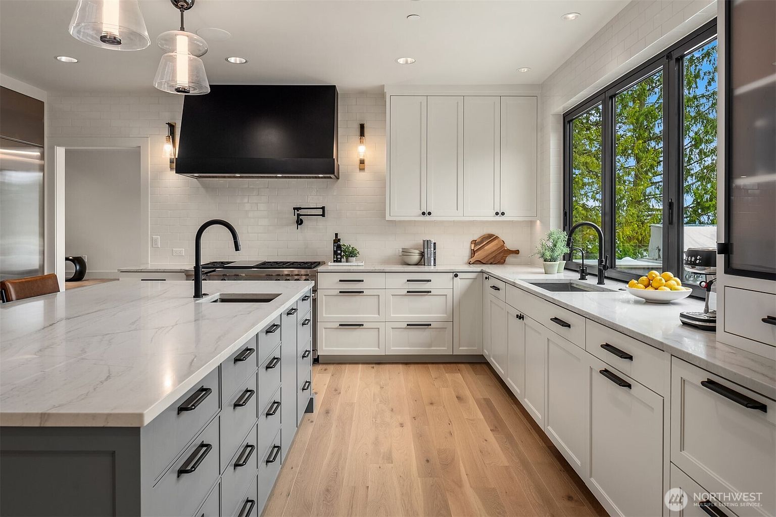 This modern, high-end kitchen features a large central island with a marble countertop, contrasting light gray cabinetry, and sleek black hardware. The space is anchored by a striking black range hood, white subway tile backsplash, and warm hardwood flooring, creating a bright and sophisticated atmosphere. Large windows allow for ample natural light, highlighting the clean lines and contemporary design of this functional culinary space.