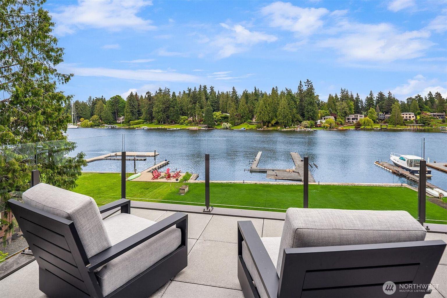 This image captures a serene, elevated view from a modern balcony overlooking a tranquil lake. Two contemporary lounge chairs with grey cushions are positioned in the foreground, facing a glass railing that provides an unobstructed perspective of the lush green lawn, private docks, and the calm water beyond. The scene exudes a peaceful, high-end waterfront lifestyle, perfect for relaxation and enjoying the scenic natural surroundings.
