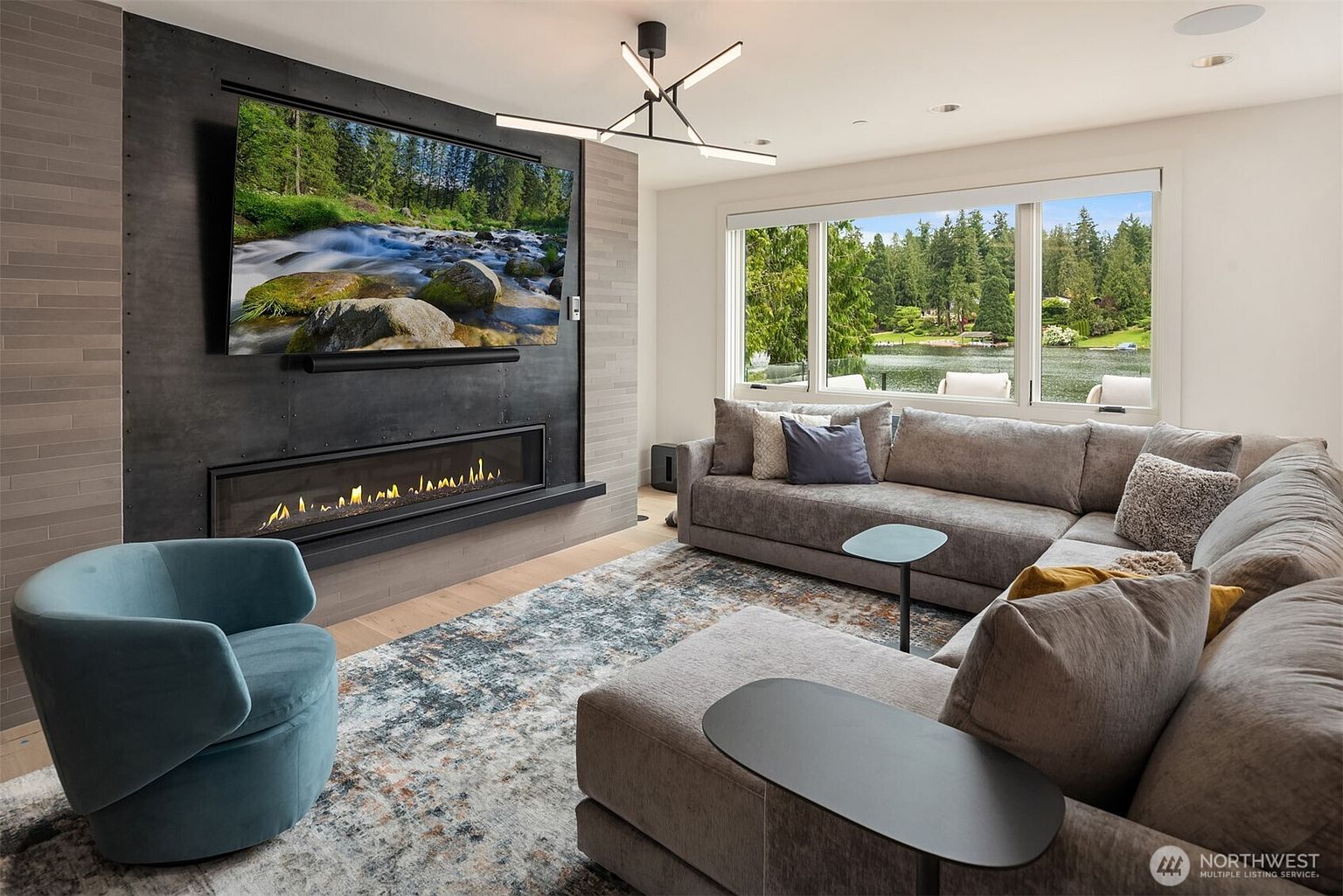 This modern living room features a striking floor-to-ceiling fireplace wall with a built-in linear fireplace and a large mounted television. A spacious, plush grey sectional sofa and a vibrant blue accent chair provide comfortable seating, while a large window offers a serene view of the lake and surrounding greenery. The room is anchored by a textured area rug and illuminated by a contemporary geometric chandelier, creating a sophisticated and inviting atmosphere.