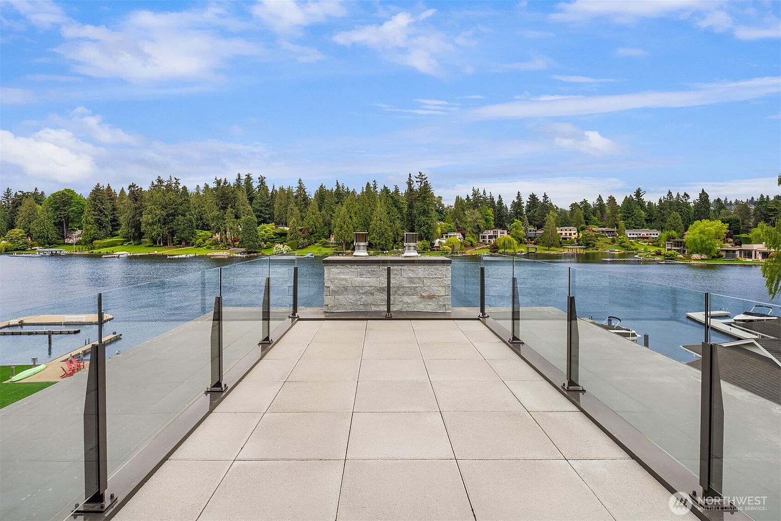 This image showcases a sleek, modern rooftop deck featuring large, light-gray stone pavers and a minimalist glass railing that offers an unobstructed, panoramic view of a serene lake. A central stone-clad chimney structure serves as a focal point, while the surrounding lush green treeline and calm water create a tranquil, high-end atmosphere. The perspective is centered and elevated, emphasizing the expansive outdoor living space and its prime waterfront location.