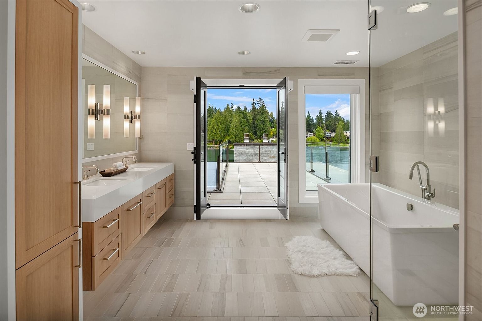 This luxurious primary bathroom features a sleek, modern design with a double vanity, a freestanding soaking tub, and a glass-enclosed shower. The room is bathed in natural light from the large windows and open French doors that lead directly to a private balcony overlooking a serene lake. The neutral color palette and clean lines create a sophisticated, spa-like atmosphere.