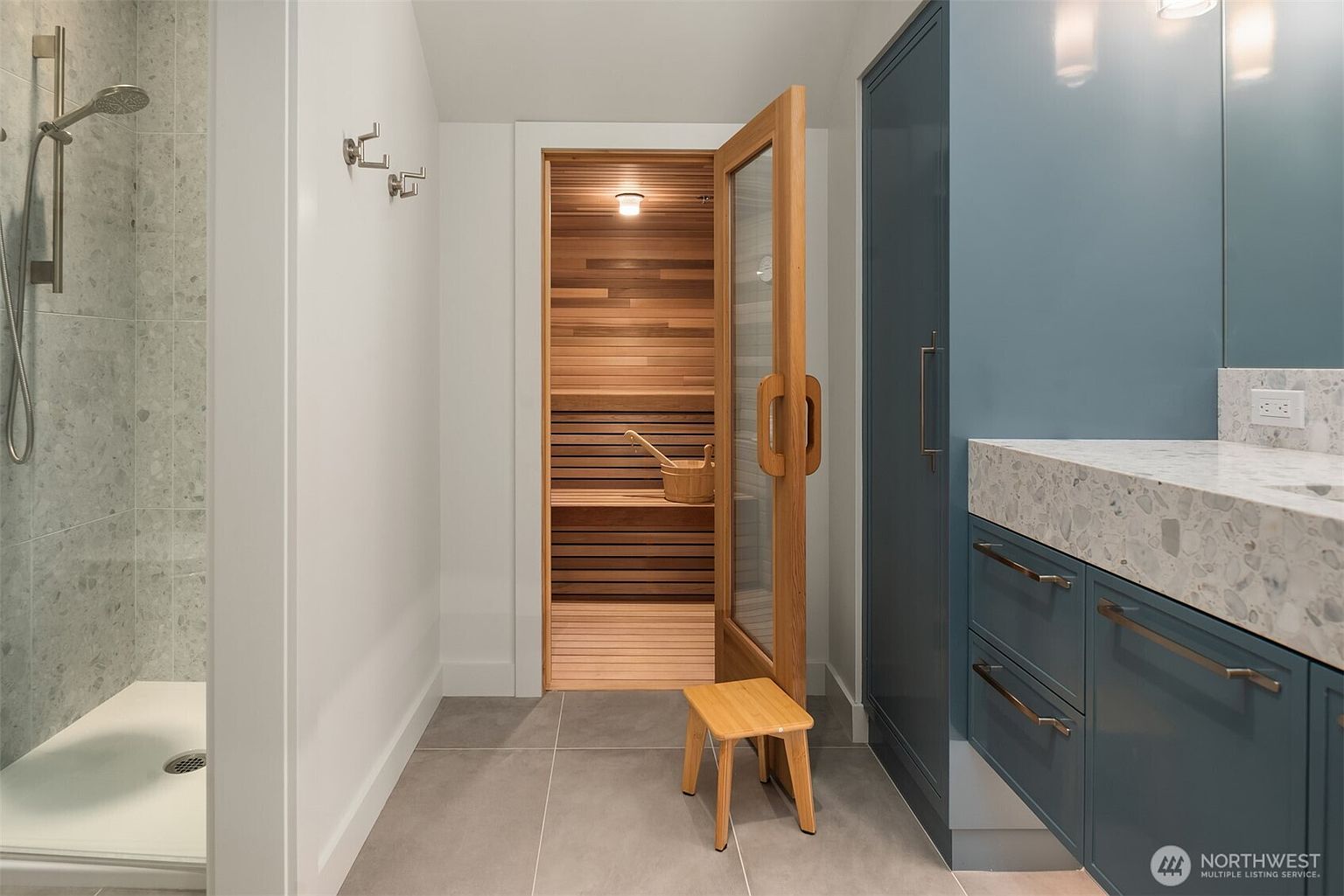 This modern, spa-inspired bathroom features a sleek walk-in shower on the left and a custom sauna with a glass door as the central focal point. To the right, a vibrant blue vanity with a stone countertop adds a pop of color, while a small wooden stool sits in the foreground. The space is finished with clean, contemporary lines and neutral tile flooring, creating a luxurious and relaxing atmosphere.