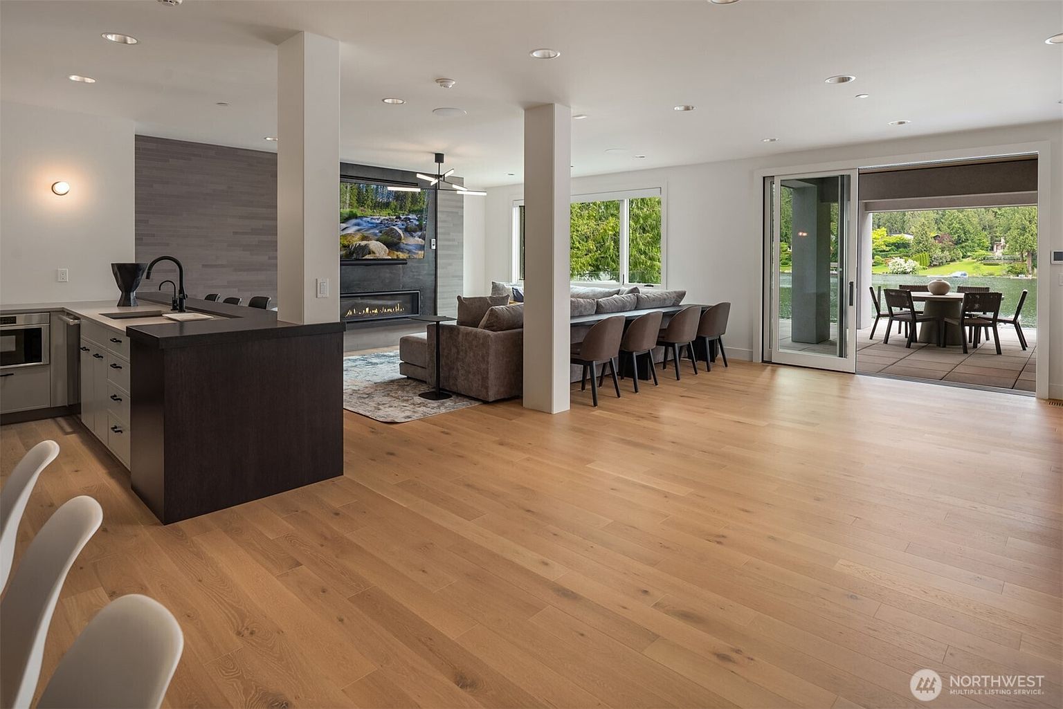 This open-concept living space seamlessly blends a modern kitchen with a cozy living area, featuring light hardwood floors and a striking dark stone fireplace wall. The room is anchored by a large grey sectional sofa and a dining table positioned behind it, while floor-to-ceiling sliding glass doors provide a direct view and access to an outdoor patio overlooking a lake. The design is contemporary and airy, characterized by clean lines, neutral tones, and abundant natural light.