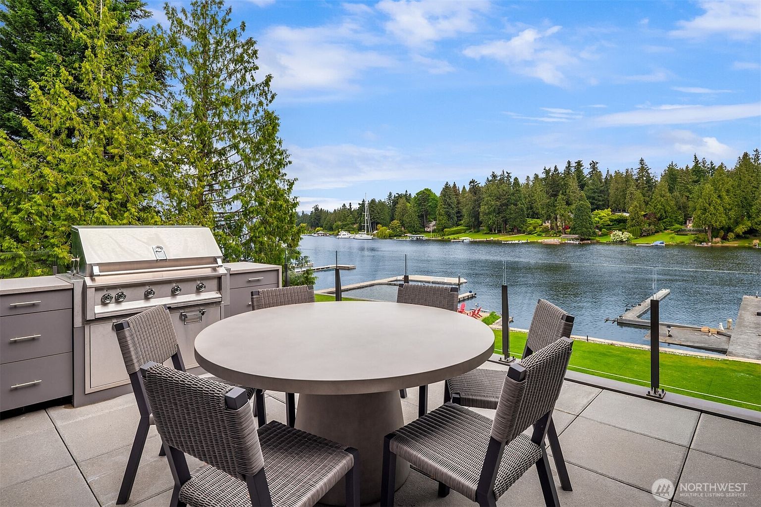 This elevated outdoor deck features a modern, circular concrete dining table surrounded by woven wicker chairs, perfect for al fresco dining. A built-in stainless steel grill station is positioned to the side, while a sleek glass railing provides an unobstructed, panoramic view of the serene lake and lush, tree-lined shoreline. The perspective captures a tranquil, high-end waterfront living space designed for relaxation and entertaining.
