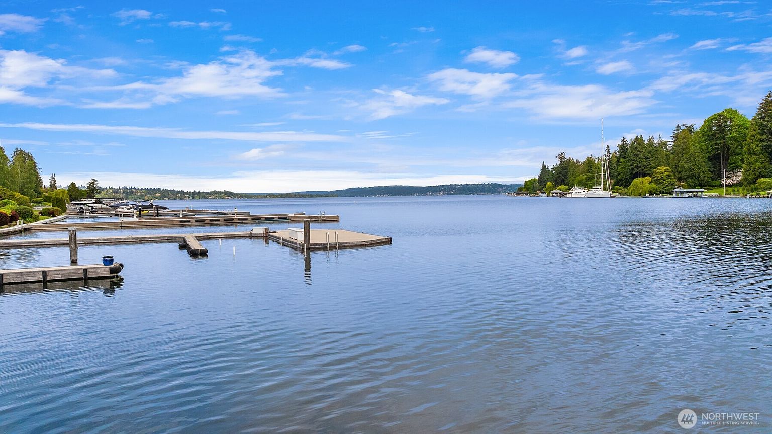 This scenic view captures a tranquil lakefront setting featuring a series of wooden boat docks extending into the calm, blue water. The perspective is wide and expansive, showcasing the lush green treeline along the distant shore under a bright, partly cloudy sky. It highlights the peaceful, recreational lifestyle associated with waterfront living.
