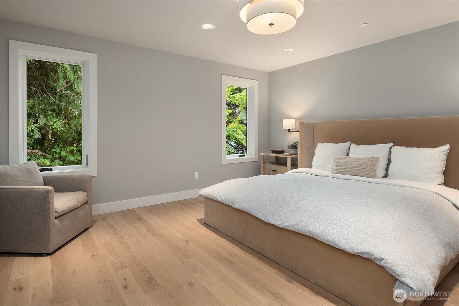 This serene primary bedroom features light hardwood flooring, soft gray walls, and a large, plush upholstered bed as the focal point. Two tall windows allow for natural light and views of the surrounding greenery, while a comfortable armchair in the corner creates a cozy reading nook. The room is illuminated by a modern, circular flush-mount ceiling light, contributing to an overall clean and tranquil aesthetic.