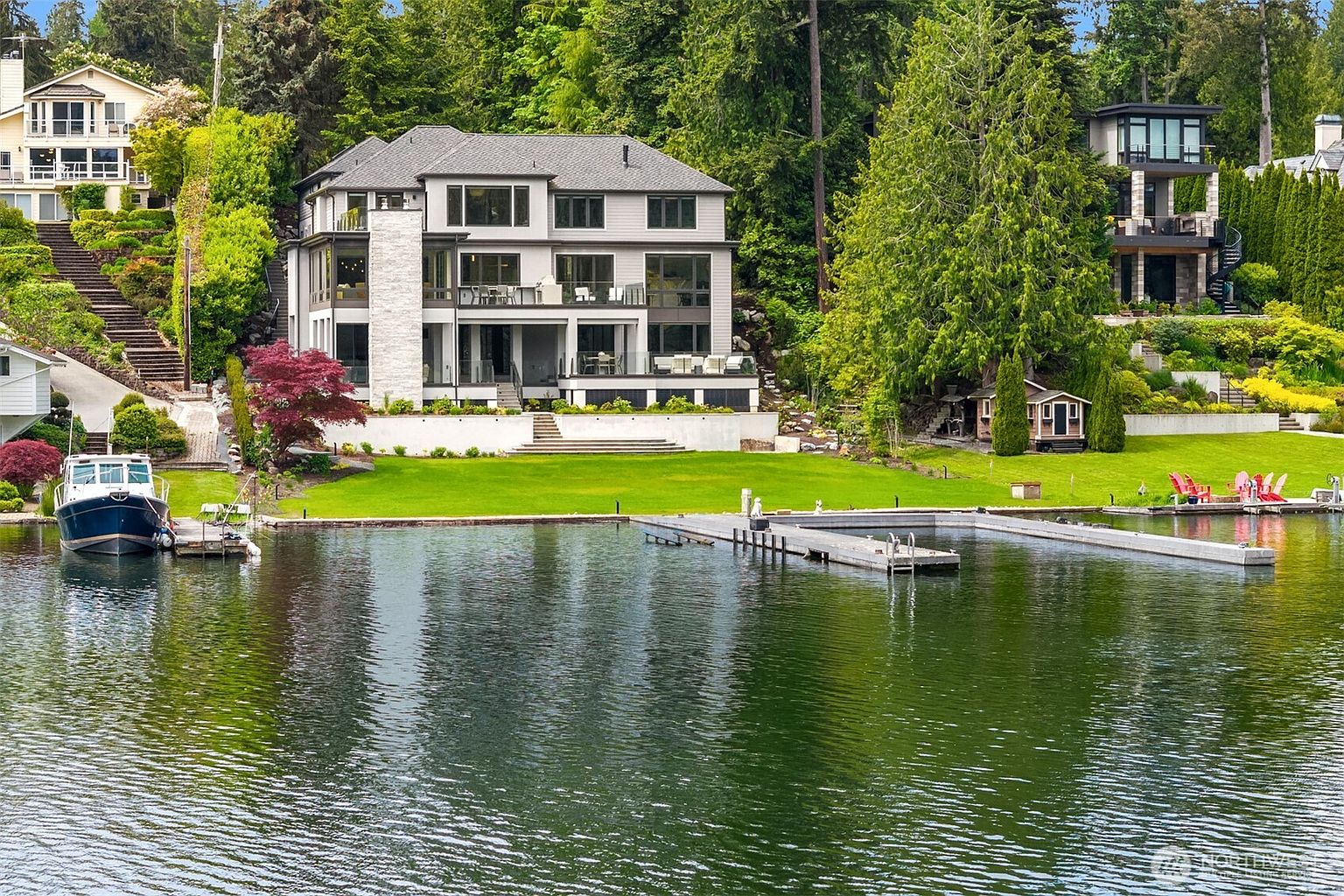 This stunning waterfront estate features a multi-level contemporary home with expansive windows and a prominent stone chimney, perfectly positioned on a lush, manicured lawn. The property includes a private dock extending into the calm lake, framed by dense evergreen trees and a gentle slope leading down to the water. The perspective is captured from the water, highlighting the seamless integration of luxury architecture with the serene natural landscape.