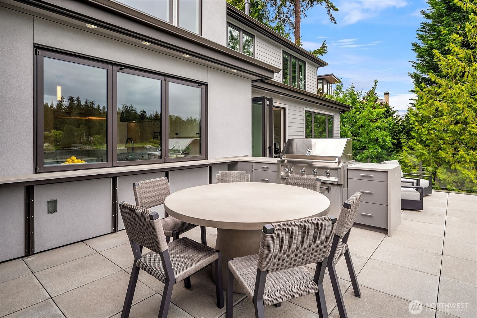 This modern outdoor patio features a sleek, minimalist design with a concrete dining table and woven chairs, perfect for al fresco entertaining. The space is equipped with a built-in stainless steel grill and countertop, seamlessly integrating with the home's contemporary gray exterior and large glass windows. The elevated perspective highlights the clean lines and functional layout of this sophisticated outdoor living area.